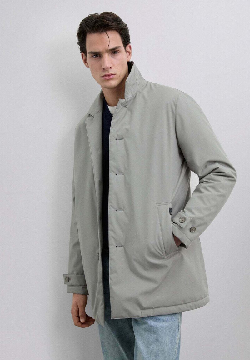 Joven vestido con un abrigo abotonado gris claro y el cuello alzado, suéter oscuro debajo y jeans azules claros, de pie contra un fondo liso.