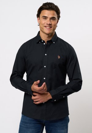 Schwarzes Langarmhemd mit knöpfbarem Kragen, weißen Knöpfen und einem kleinen gestickten Logo auf der Brust, kombiniert mit blauen Jeans.