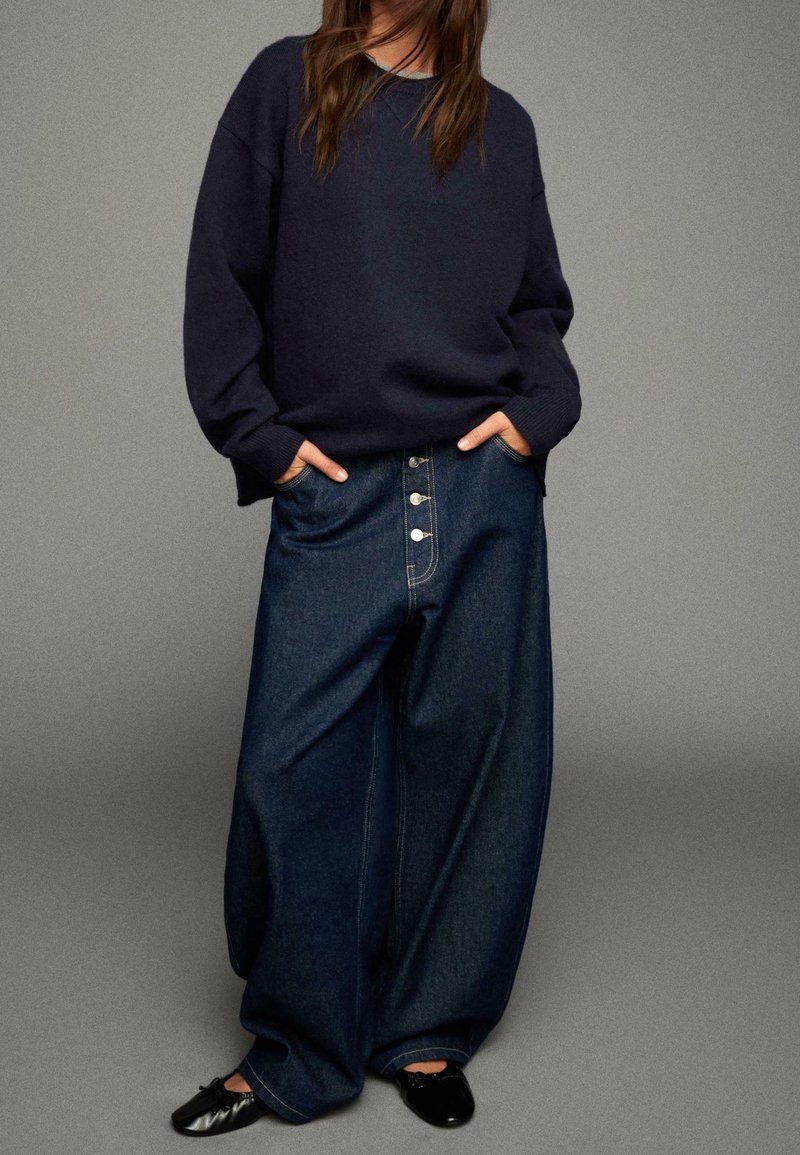 Personne portant un pull ample bleu marine foncé, un jean large taille haute en denim foncé avec des boutons, et des ballerines noires, debout les mains dans les poches.