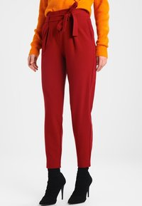 Röda tapered byxor med hög midja, veck och ett självknutet bälte. Tillverkade av ett slätt material, ihop med svarta klackstövlar.