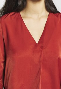 Blouse rouge avec un col en V profond, une texture lisse et un design légèrement drapé. Des accents cousus près du col ajoutent un détail subtil.