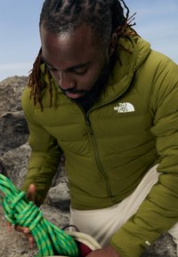 Giacca a vento verde con chiusura a zip e logo sul petto, indossata da una persona che tiene una corda da arrampicata verde, in piedi vicino a delle rocce.