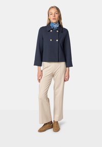 Navy cropped double-breasted jacket with gold buttons, paired with beige flared pants and tan suede sneakers. Blue patterned scarf around neck.