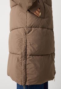 Manteau matelassé marron avec un design matelassé, comportant une poche latérale et un détail de fermeture éclair en bas. Le matériau semble léger et doux.