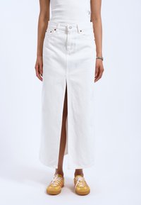 Gonna maxi in denim bianco con uno spacco frontale, caratterizzata da un taglio dritto e design a cinque tasche. Abbinata a sneakers gialle con lacci bianchi.