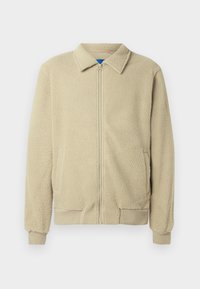 Veste en polaire beige avec fermeture éclair frontale, col montant, deux poches latérales, poignets et ourlet côtelés. La texture semble douce et confortable.