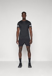 Schwarzes Sport-T-Shirt mit weißem "BORG"-Logo auf den Ärmeln, kombiniert mit schwarzen Shorts und schwarzen Sneakers, vor einem neutralen Hintergrund stehend.