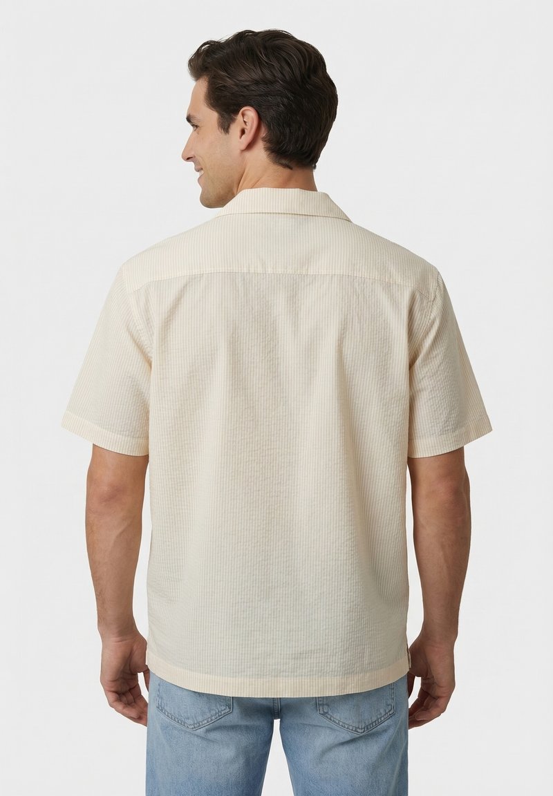 Homme portant une chemise beige texturée à manches courtes et un jean bleu clair, de dos devant un fond blanc uni.