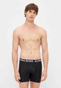 Noor nägu heleda naha ja lokkis pruunide juustega noormees, kes kannab musti Björn Borg boxer-aluspesu, seisab tasase valge tausta ees.