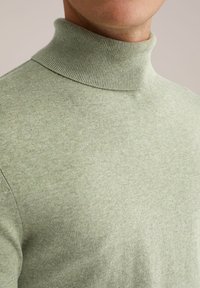 Hellgrüner Rollkragenpullover aus weichem Strickstoff, mit geripptem Kragen und glatter Textur. Nahaufnahme des Oberteils.