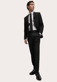 Zwart pak met een nauwsluitende pasvorm, wit overhemd en zwarte stropdas. Glanzende zwarte schoenen en klassieke designelementen zorgen voor een formele uitstraling.