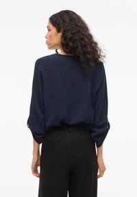 Μπλούζα σε ναυτικό μπλε με υφή, άνετη εφαρμογή και γυριστά μανίκια, συνδυασμένη με μαύρο παντελόνι με ελαστική μέση. Μινιμα λιστικός σχεδιασμός, χωρίς ορατά σχέδια.