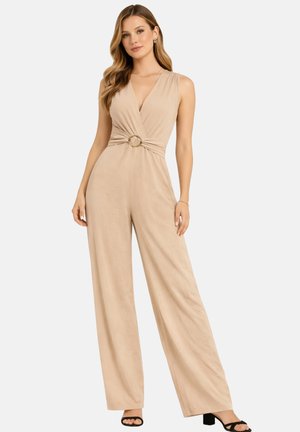Kvinde iført ærmeløs beige jumpsuit med vide ben og dyb V-hals med guldringdetalje ved taljen, kombineret med sorte åbne hæle.