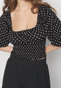 Svart blus med vita prickar, med fyrkantig halsringning, puffärmar och en smockad midja för en figursydd form.