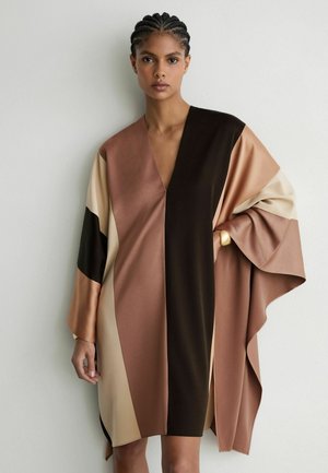 Femme aux cheveux tressés portant une robe ample à blocs de couleurs dans des tons marron, beige et noir, debout devant un mur blanc uni.