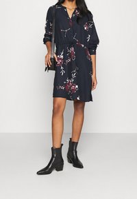 Marine jurk met bloemenpatroon, knoopsluiting aan de voorkant, lange mouwen en een striktaille. Draag het met zwarte enkelboots en een kleine crossbody tas.