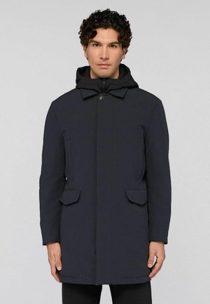 Homme portant un manteau d'hiver à capuche bleu marine foncé avec des poches avant, fermeture à boutons et à glissière, se tenant devant un fond gris clair uniforme.