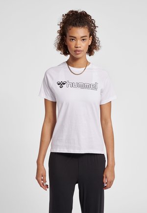 Femme aux cheveux bouclés portant un t-shirt blanc à manches courtes Hummel et un pantalon noir, debout devant un fond clair uni.