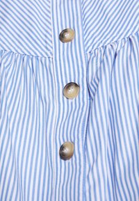 Tessuto di cotone a righe blu e bianche con linee verticali, caratterizzato da grandi bottoni marroni lungo la parte anteriore e dettagli arricciati nella parte superiore.