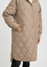 Manteau matelassé beige clair avec une fermeture à boutons, un motif en losanges et des poches latérales. Tissu doux avec une coupe décontractée et un ourlet arrondi.