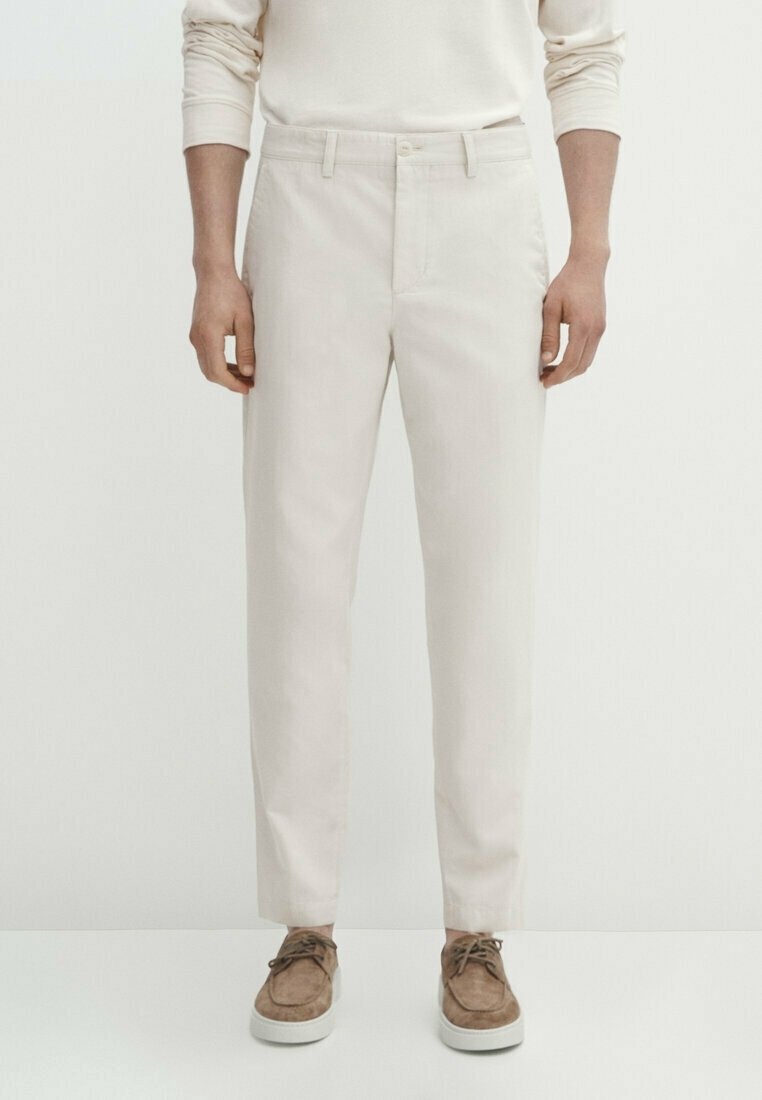 Homme portant un pantalon beige, une chemise beige à manches longues et des chaussures en daim marron avec des semelles blanches, debout sur un fond clair uni.