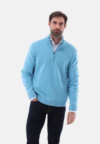 Hellblaues Zip-Kragen-Sweatshirt mit gerippten Ärmelabschlüssen und Saum, aus weichem Material, über einem weißen Hemd mit Kragen getragen, kombiniert mit dunklen Jeans.