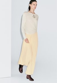 Vrouw die loopt in een crèmekleurige top met lange mouwen, beige geplooide maxi-rok, bruine enkellaarsjes en een ronde hangersketting.