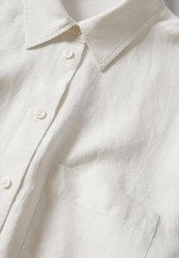Chemise en lin blanc avec col boutonné, poche avant et texture subtile. Fermeture avec des boutons ronds blancs sur le devant.