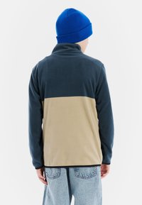 Fleece-Jacke mit einem oberen Teil in Marineblau und einem unteren Teil in Tan. Verfügt über einen Kragen, lange Ärmel und einen lässigen Schnitt. Begleitet von einer blauen Mütze.