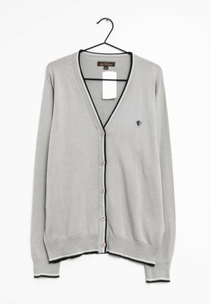 Hellgraue Strickjacke mit Knöpfen, schwarzem und weißem Besatz, langen Ärmeln und kleinem Schild-Emblem auf der linken Brust, aufgehängt an einem schwarzen Bügel.