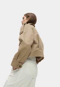 Kort beige jack met knoopmanchetten en gestructureerde schouders, gedragen over lichte broek. Glad, matte stof met minimale details.