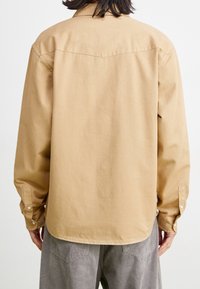 Hiekanvärinen pitkähiha-pellavapaita, jossa on rento istuvuus, napilliset kalvosimet ja takaosassa ylaosaommelma. Sileä pinta ja vähäiset yksityiskohdat.