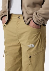 Beige outdoor pants with a relaxed fit, featuring zippered pockets and logo detail. The fabric is lightweight and textured for comfort.