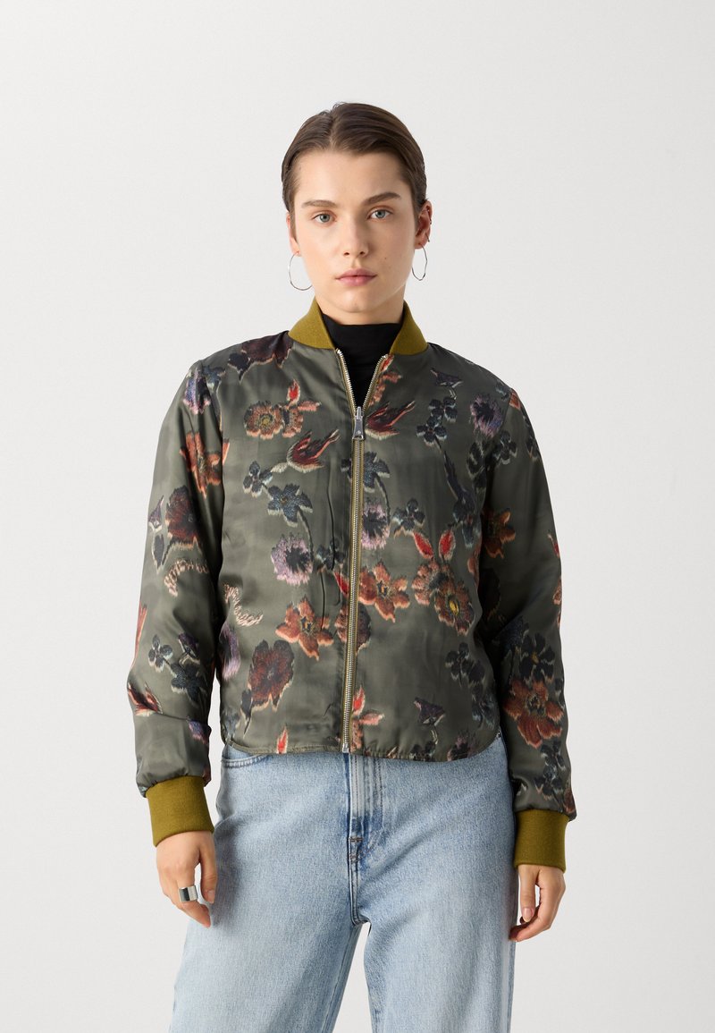 Veste bomber florale en satin vert olive, avec une fermeture éclair sur le devant, un col et des poignets côtelés. Motif floral dans plusieurs couleurs. Portée sur un jean.
