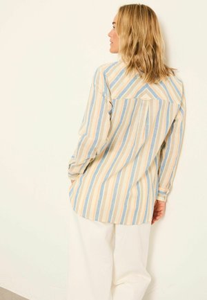 Woman with shoulder-length blonde hair wears a long-sleeve, striped shirt in beige, blue, and white, paired with loose white pants.