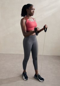 Model fitness trzymający taśmy oporowe, ubrany w czerwoną cropped top i szare legginsy. Czarne sneakersy z białymi akcentami. Jasne tło.