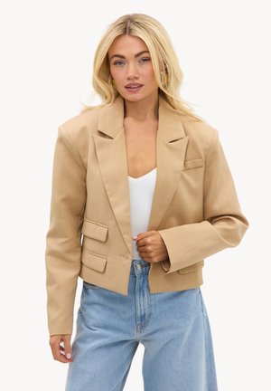Femme aux cheveux blonds portant un blazer beige court sur un top blanc et un jean bleu clair, regardant devant avec une main près du blazer.