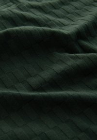 Tejido texturizado de color verde oscuro con un patrón de cuadrícula, que presenta cuadrados elevados y planos alternados. Material suave y flexible con un sutil brillo.
