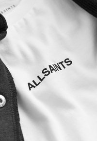 Čierna bavlnená látka s textúrovaným golierom sa stretáva s bielou košeľou. Logo "ALLSAINTS" je tučne vytlačené čiernym písmom a je umiestnené v strede bielej oblasti.