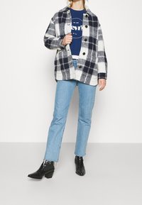 Giacca in misto lana a quadri blu navy e bianco, con grandi bottoni neri, indossata sopra una felpa blu Levi's, abbinata a jeans azzurri e stivali neri con tacco.