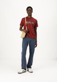 Camiseta roja de manga corta con el gráfico "REPLAY", vaqueros azules, bolso cruzado beige y zapatillas en blanco y negro; de pie sobre un fondo blanco.