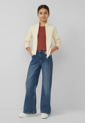 Młoda dziewczyna stojąca z rękami w kieszeniach, ubrana w beżową kurtkę na zamek, rdzawy T-shirt, szerokie niebieskie dżinsy i białe trampki.