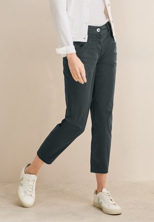 Vrouw met cropped donkere broek, witte sneakers en een wit jack, lopend tegen een beige achtergrond.