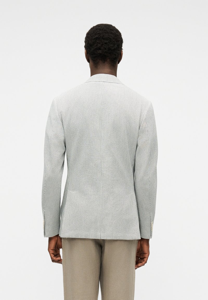 Uomo che indossa un blazer grigio chiaro con trama e pantaloni beige, visto da dietro su uno sfondo semplice.