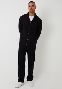 Cárdigan negro de punto con cuello chal, que presenta un patrón de trenzas y grandes botones. Camiseta blanca debajo, pantalones negros y zapatos blancos.
