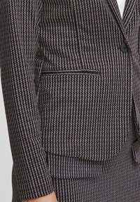 Črno-bela blazer jakna s patterjem, ki ima prilegajočo zasnovo, enojno gumbasto zapiranje in stranski žep za dodatne podrobnosti.