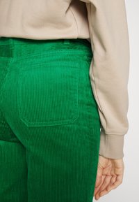 Un pantalon en velours côtelé vert présente des côtes verticales prononcées, des poches arrière et une coupe ajustée, associé à un haut beige à manches longues.