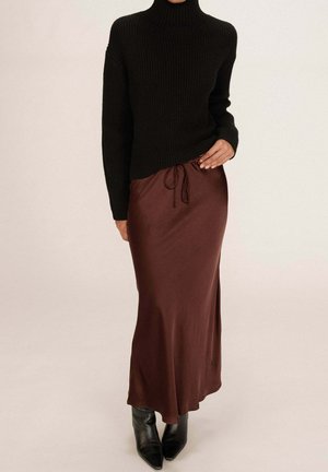 Femme portant un pull à col roulé noir en maille côtelée, une jupe midi en satin brun avec taille à cordon et des bottes noires à bout pointu.