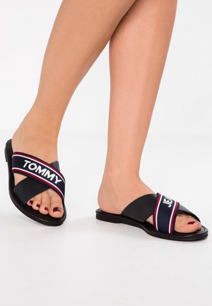 Fødder iført sorte slip-on sandaler med krydsede stropper mærket ""TOMMY JEANS"" i hvidt på marineblåt med rød og hvid kant.