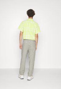 Man with curly hair wearing a short-sleeve lime green plaid shirt, black-and-white checkered pants, and white sneakers, standing against a white wall.